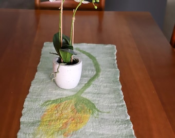 May include: A light green felt table runner with a yellow and green floral design. A white ceramic pot with an orchid plant sits on the runner. The runner is on a wooden table.