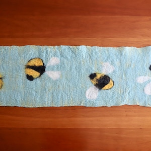 May include: A light blue felt table runner with four yellow and black bee designs. The bees are flying with white wings.