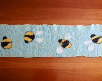 May include: A light blue felt table runner with four yellow and black bee designs. The bees are flying with white wings.