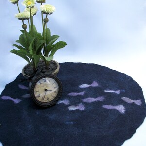 May include: A dark blue felt rug with a design of light purple fish. A small potted plant with white flowers and a clock are on top of the rug.