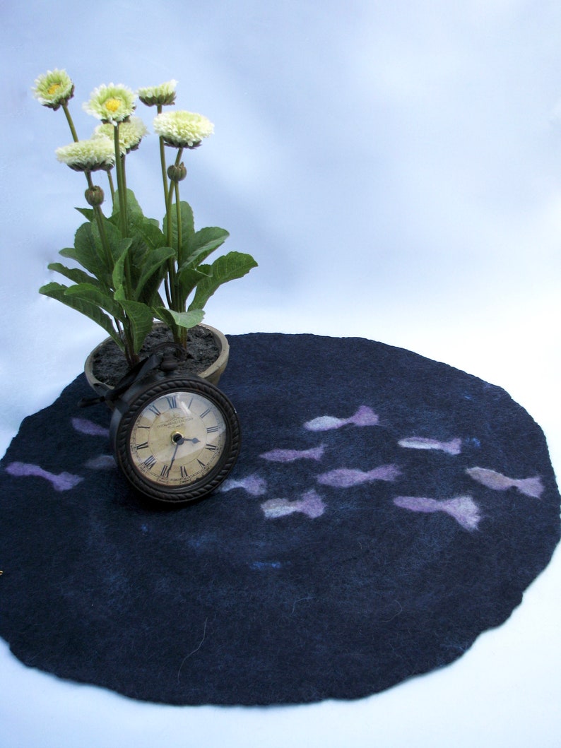 May include: A dark blue felt rug with a design of light purple fish. A small potted plant with white flowers and a clock are on top of the rug.