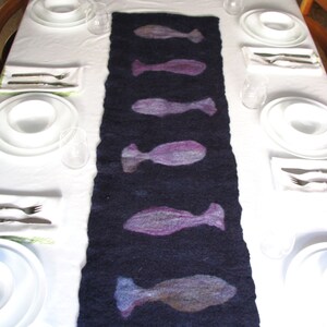 May include: A dark blue table runner with a repeating pattern of purple and gray fish silhouettes. The runner is laid out on a white tablecloth with place settings on either side.