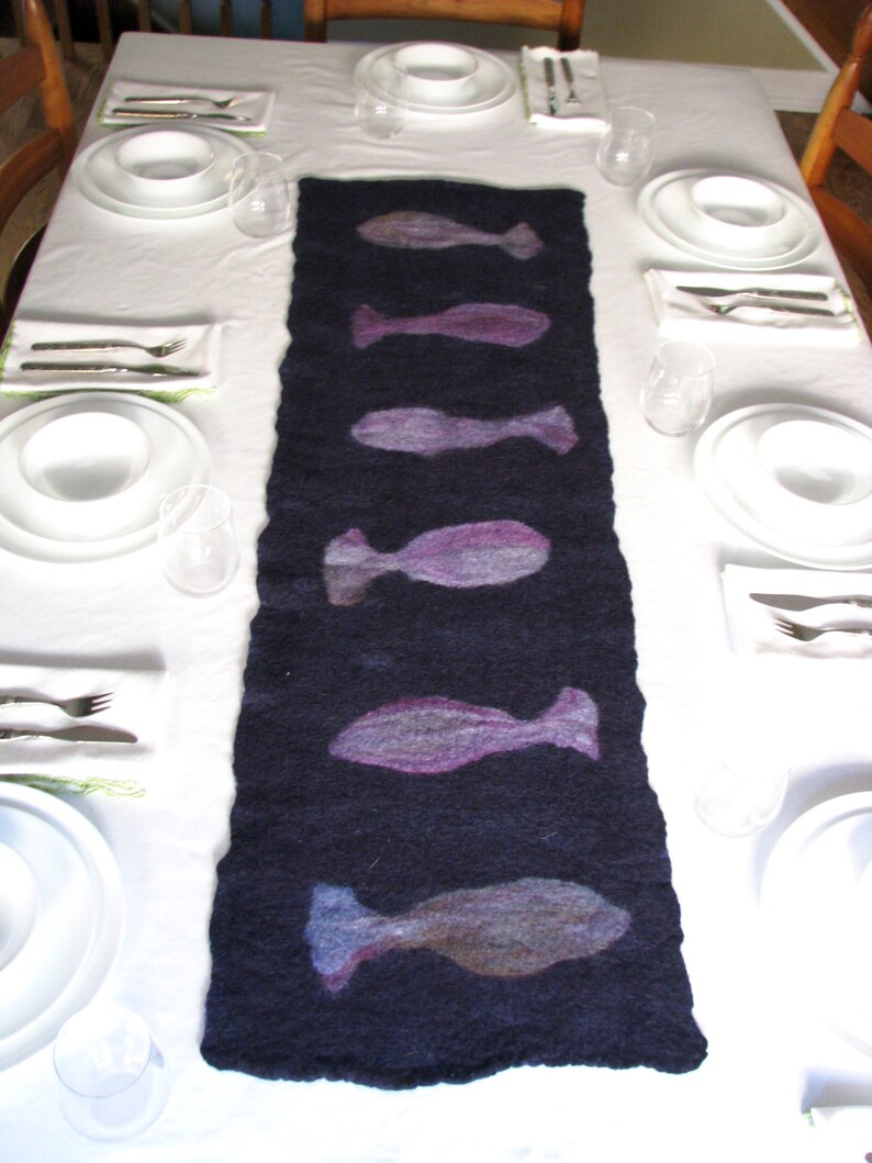 May include: A dark blue table runner with a repeating pattern of purple and gray fish silhouettes. The runner is laid out on a white tablecloth with place settings on either side.