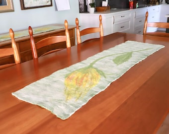 May include: A handmade, light green table runner with a large yellow and orange flower design. The runner is placed on a wooden table, with wooden chairs surrounding it. The runner adds a decorative touch to the dining area.