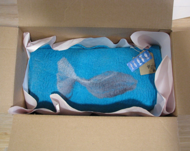 May include: A blue felt wall hanging with a white fish design. The hanging is packaged in a cardboard box with pink tissue paper.