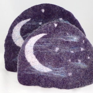 May include: Two purple felt moon and stars ornaments. The ornaments are shaped like half circles and have a white crescent moon and white stars on them.