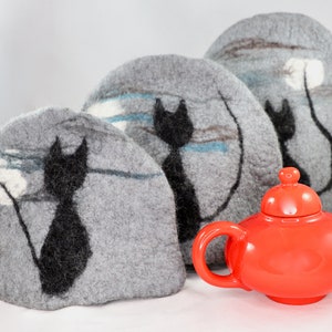 May include: Three grey felt tea cosy covers with black cat silhouettes and white accents. The covers are arranged in a row, with the largest at the back and the smallest at the front. A red ceramic teapot sits in front of the covers.