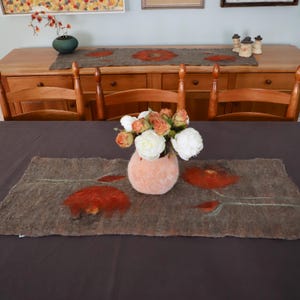 May include: A gray felt table runner with a floral design featuring red poppies. The runner is on a dark brown table with a vase of white and peach flowers in the center.