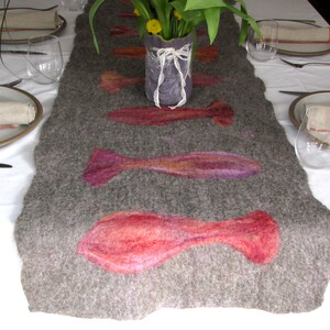 May include: A gray felt table runner with a decorative design of pink, purple, and orange fish. The runner is on a white tablecloth with a vase of flowers in the center.