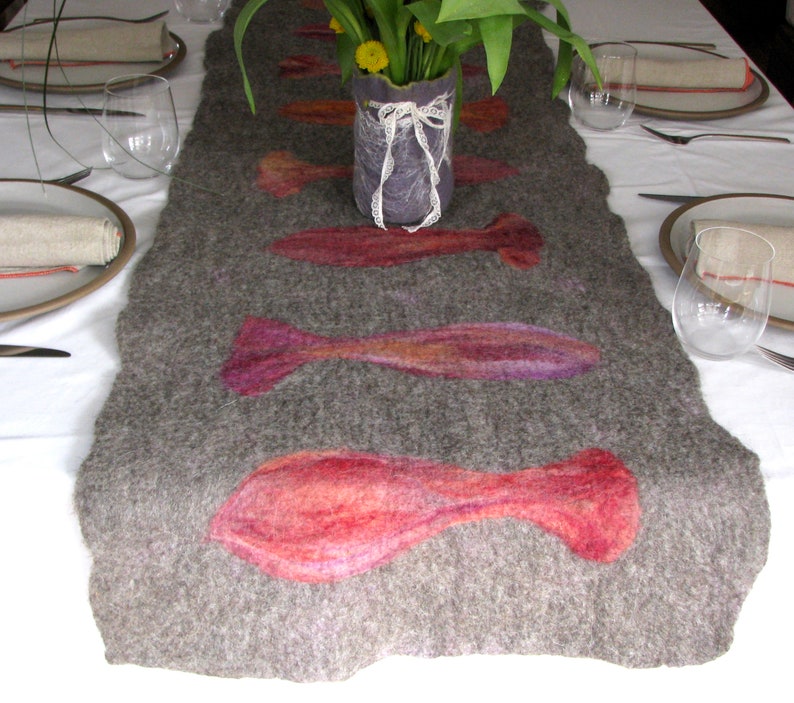 May include: A gray felt table runner with a decorative design of pink, purple, and orange fish. The runner is on a white tablecloth with a vase of flowers in the center.