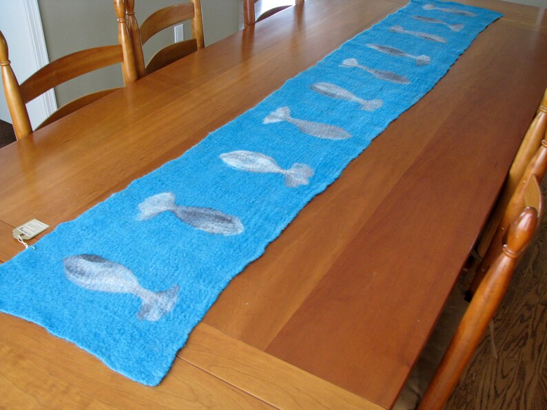 May include: A blue table runner with a white fish design. The runner is on a wooden table.
