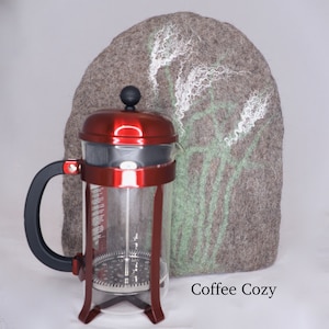 May include: A red and clear glass French press coffee maker with a grey felt coffee cosy with a green and white floral design. The cosy has the text "Coffee Cozy" printed on it.