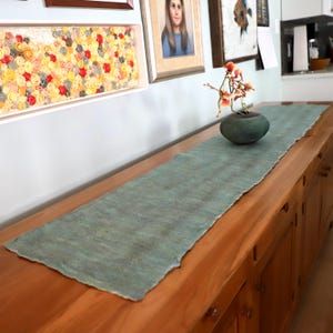 Peut inclure: Une longue nappe de table rectangulaire de couleur sarcelle avec un bord brut et irrégulier. Elle est posée sur un buffet en bois. Un petit vase vert foncé avec une plante est posé sur la nappe. Des œuvres d'art et des photos encadrées sont au mur.