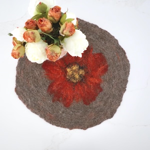 May include: A round, grey felt coaster with a red flower design in the centre. The flower has a yellow centre and orange petals.