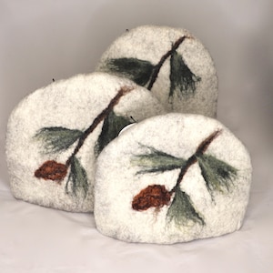 May include: Three off-white felt tea cosies with a pine cone and branch design. The design features a brown pine cone, green needles, and a brown branch. The tea cosies are arranged in a layered composition.