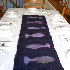 May include: A dark blue table runner with a white background. The runner has a design of five purple fish on it. The table is set with white plates, bowls, and silverware.