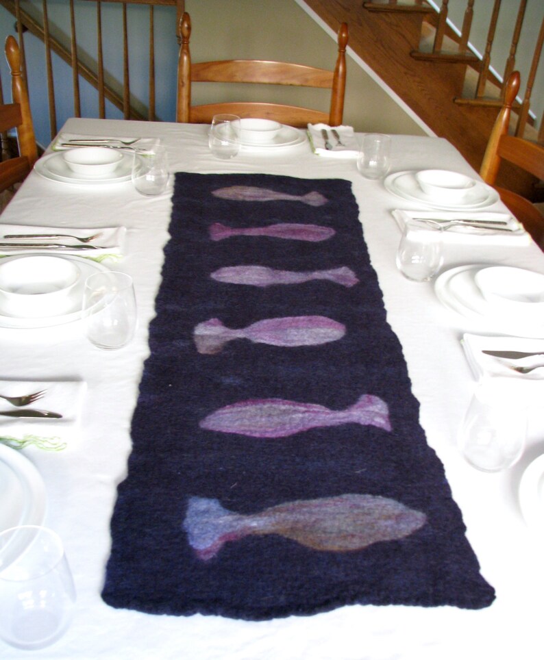 May include: A dark blue table runner with a white background. The runner has a design of five purple fish on it. The table is set with white plates, bowls, and silverware.