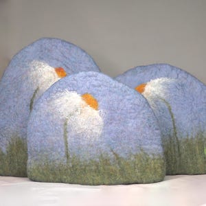 May include: Three felted sculptures in a soft, muted color palette. Each piece features a light blue arch with a white and orange floral design. The base of each sculpture has a green, grassy texture.