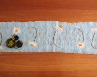 May include: A light blue felt table runner with white felt flowers and green felt stems. Three small black and brown woven baskets with green moss are on the runner.