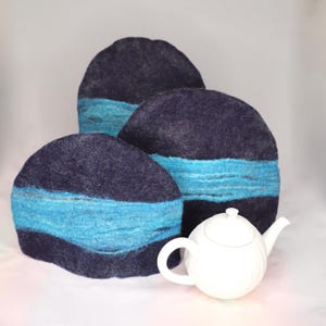 May include: Three navy blue tea cosies with a turquoise band, and a white teapot. The tea cosies are round and made of a felted material. The teapot is white with a curved handle and spout. The background is white.