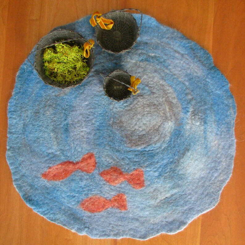 May include: A blue felt rug with a swirling pattern, resembling a pond, with three small woven baskets and three orange fish shapes.