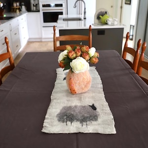 May include: A white and orange felted vase with a bouquet of peach and white roses sits on a white and grey sheep-themed table runner on a dark grey table.