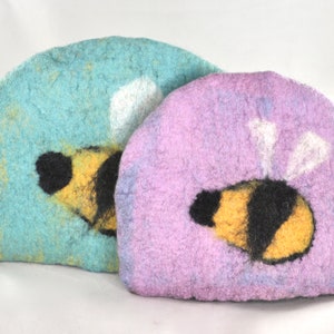 May include: Two felted wool tea cozy covers with bee designs. One is light blue with a yellow bee and the other is purple with a yellow bee.