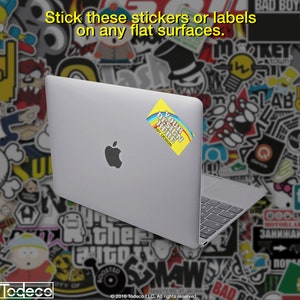 Puede incluir: Un ordenador port&aacute;til plateado con una pegatina amarilla que dice "Your Design Here" con un dise&ntilde;o de arco&iacute;ris. El port&aacute;til est&aacute; sobre un fondo de pegatinas de colores.