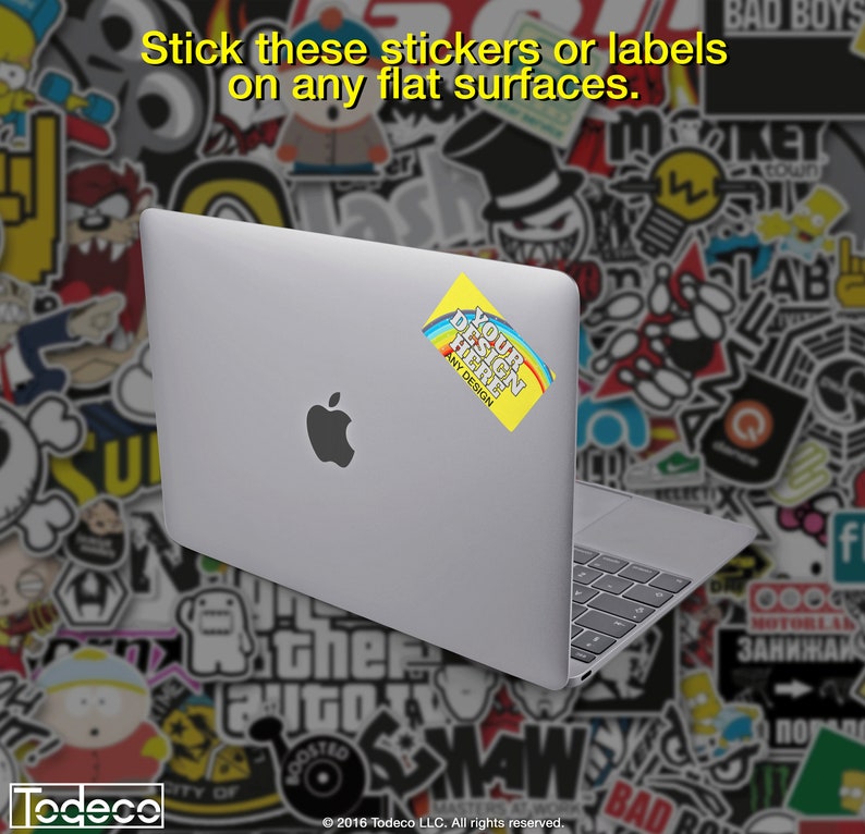 Puede incluir: Un ordenador port&aacute;til plateado con una pegatina amarilla que dice "Your Design Here" con un dise&ntilde;o de arco&iacute;ris. El port&aacute;til est&aacute; sobre un fondo de pegatinas de colores.