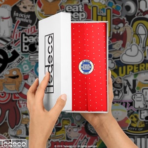 Puede incluir: Una caja blanca con la palabra "Todeco" impresa en el lateral est&aacute; abierta, revelando un papel de regalo rojo con lunares blancos. El papel de regalo tiene un c&iacute;rculo azul con estrellas blancas y el texto "ANY DESIGN YOUR DESIGN HERE ANY COT" impreso en &eacute;l.