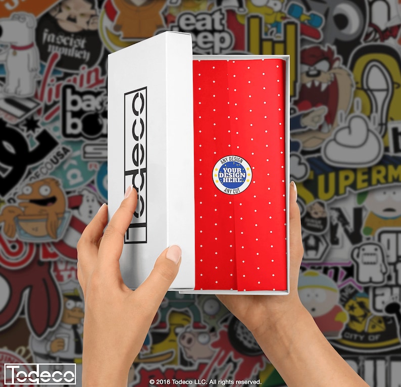 Puede incluir: Una caja blanca con la palabra "Todeco" impresa en el lateral est&aacute; abierta, revelando un papel de regalo rojo con lunares blancos. El papel de regalo tiene un c&iacute;rculo azul con estrellas blancas y el texto "ANY DESIGN YOUR DESIGN HERE ANY COT" impreso en &eacute;l.