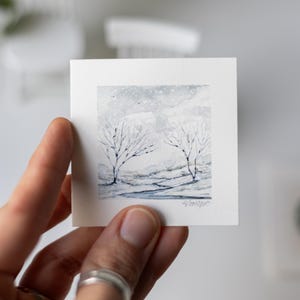 May include: A small, square watercolor painting depicting a winter landscape with two bare trees under a snowy sky. The artwork is primarily in shades of gray and white, with delicate brushstrokes. The painting is held in a hand.