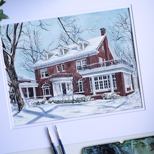 Op de afbeelding: Waterverfschilderij van een bakstenen huis in de sneeuw. Het huis heeft een witte veranda en een zwart balkon. Kale bomen omringen het huis en de lucht is lichtblauw. Penselen en een aquarelpalet staan op de voorgrond.