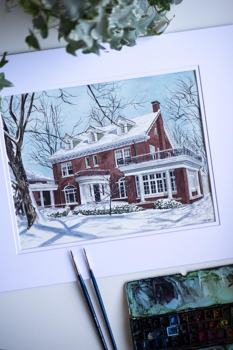 Op de afbeelding: Waterverfschilderij van een bakstenen huis in de sneeuw. Het huis heeft een witte veranda en een zwart balkon. Kale bomen omringen het huis en de lucht is lichtblauw. Penselen en een aquarelpalet staan op de voorgrond.