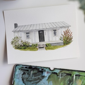 Op de afbeelding: Waterverfschilderij van een wit huis met een grijs dak, zwarte deur en het nummer 949 boven de deur. Het huis is omgeven door groene struiken en gras. Een aquarelpalet staat op de voorgrond.