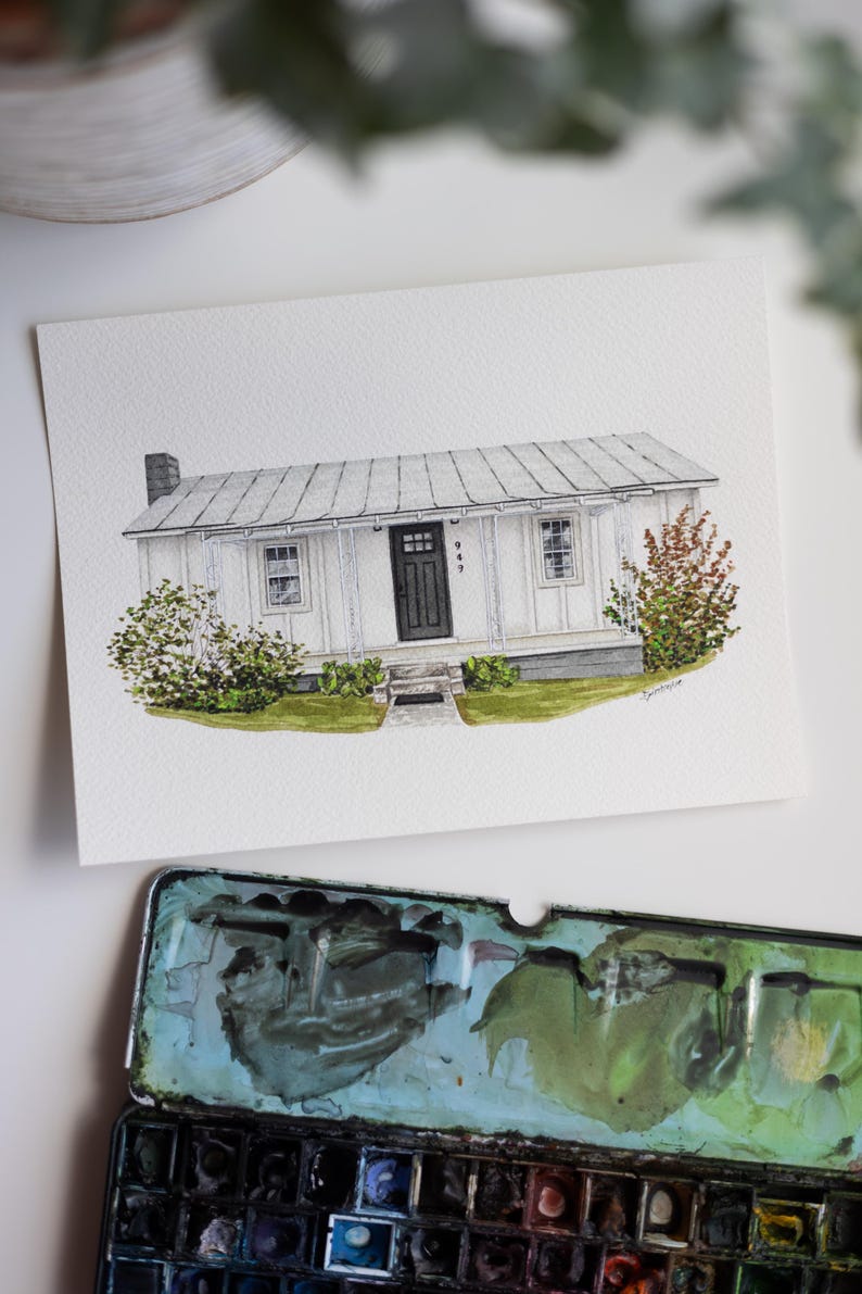 Op de afbeelding: Waterverfschilderij van een wit huis met een grijs dak, zwarte deur en het nummer 949 boven de deur. Het huis is omgeven door groene struiken en gras. Een aquarelpalet staat op de voorgrond.