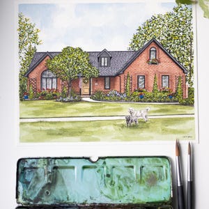 Op de afbeelding: Aquarel schilderij van een bakstenen huis met een zwart dak, een groen gazon en twee honden. De schildering bevat een Amerikaanse vlag en is omgeven door bomen en bloemen. Een aquarelverfpalet en penselen staan op de voorgrond.