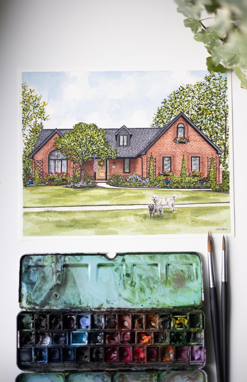 Op de afbeelding: Aquarel schilderij van een bakstenen huis met een zwart dak, een groen gazon en twee honden. De schildering bevat een Amerikaanse vlag en is omgeven door bomen en bloemen. Een aquarelverfpalet en penselen staan op de voorgrond.