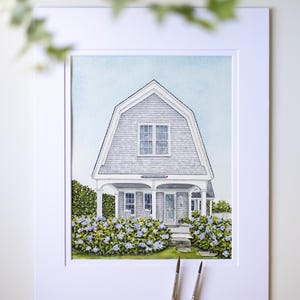 Op de afbeelding: Aquarel schilderij van een grijs huis met een kenmerkend dak, wit omlijst. Het huis heeft een veranda met twee stoelen en is omgeven door blauw en groen gebladerte. Twee penselen staan op de voorgrond.