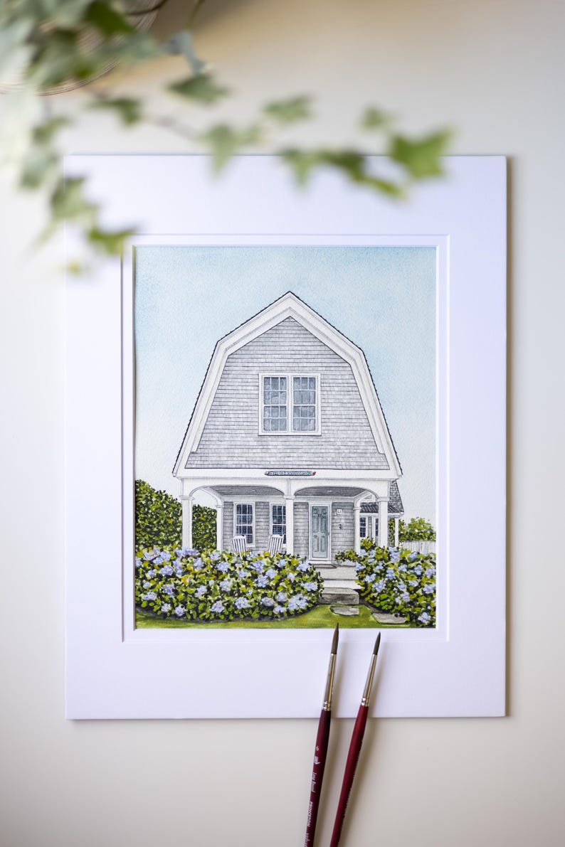 Op de afbeelding: Aquarel schilderij van een grijs huis met een kenmerkend dak, wit omlijst. Het huis heeft een veranda met twee stoelen en is omgeven door blauw en groen gebladerte. Twee penselen staan op de voorgrond.