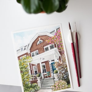 Op de afbeelding: Waterverfschilderij van een bakstenen huis met een witte veranda en blauwe deur, omgeven door groen en bloemen. Twee penselen met rode handvatten liggen naast het kunstwerk. Een potplant staat op de achtergrond.