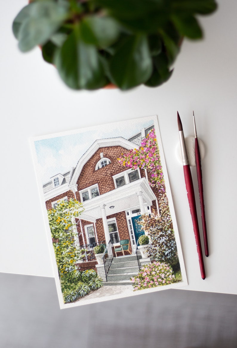Op de afbeelding: Waterverfschilderij van een bakstenen huis met een witte veranda en blauwe deur, omgeven door groen en bloemen. Twee penselen met rode handvatten liggen naast het kunstwerk. Een potplant staat op de achtergrond.