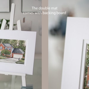 Op de afbeelding: Een ingelijste aquarel van een huis met een rode bakstenen buitenkant, tentoongesteld op een witte ezel. De dubbele passe-partout wordt geleverd met een achterbord. De schildering staat tegen een achtergrond van groene bomen en een blauwe lucht.