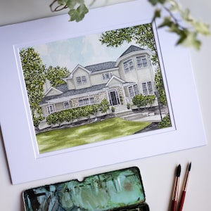 Op de afbeelding: Aquarel schilderij van een huis in een witte passe-partout. Het huis heeft een lichtgekleurde buitenkant, een stenen gevel en een donker dak. Groene bomen en struiken omringen het huis. Een palet met aquarelverf en twee penselen staan op de voorgrond.