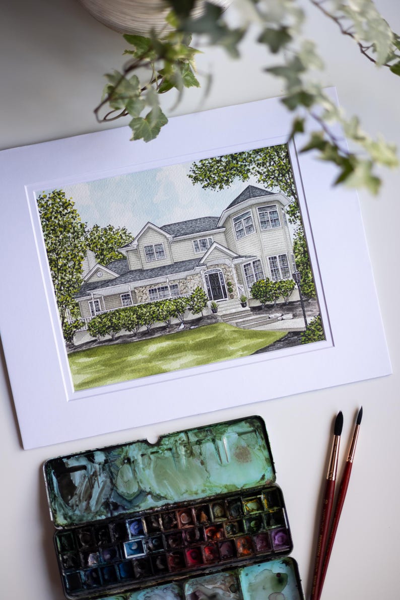 Op de afbeelding: Aquarel schilderij van een huis in een witte passe-partout. Het huis heeft een lichtgekleurde buitenkant, een stenen gevel en een donker dak. Groene bomen en struiken omringen het huis. Een palet met aquarelverf en twee penselen staan op de voorgrond.