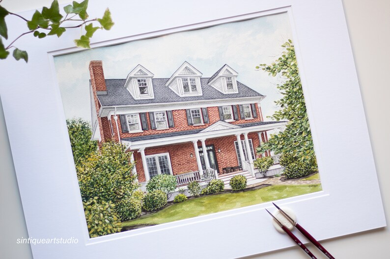 Op de afbeelding: Aquarel schilderij van een bakstenen huis met twee verdiepingen met een witte veranda en zwarte luiken. Het huis heeft een donkergrijs dak en drie dakkapellen. Groene struiken en bomen omringen het huis. Het kunstwerk is ingelijst.