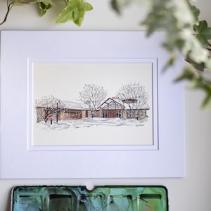 Op de afbeelding: Aquarel schilderij van een gebouw in een besneeuwd landschap, omlijst in een witte passe-partout. Het gebouw heeft een rood dak en is omgeven door kale bomen. Een penseel en een aquarelpalet staan op de voorgrond.