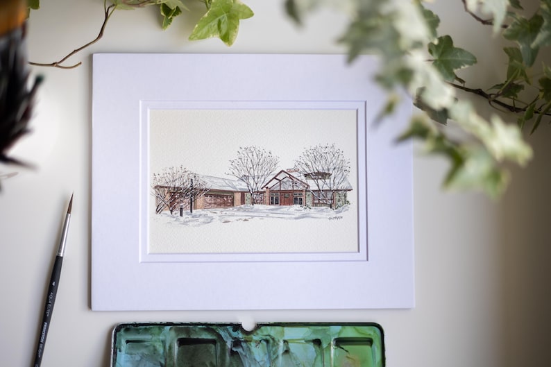Op de afbeelding: Aquarel schilderij van een gebouw in een besneeuwd landschap, omlijst in een witte passe-partout. Het gebouw heeft een rood dak en is omgeven door kale bomen. Een penseel en een aquarelpalet staan op de voorgrond.