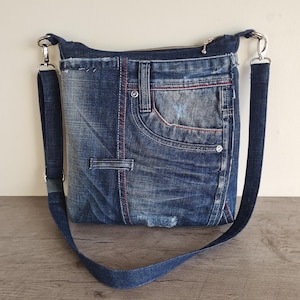 Könnte beinhalten: Eine Jeans-Umhängetasche mit Reißverschluss und verstellbarem Riemen. Die Tasche besteht aus recycelten blauen Jeans und verfügt über die originalen Taschen und rote Nahtdetails. Die Tasche hat silberfarbene Hardware.