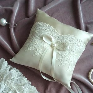 May include: A white satin ring pillow with delicate lace trim and a satin ribbon tied in a bow. The pillow is decorated with small pearl accents.